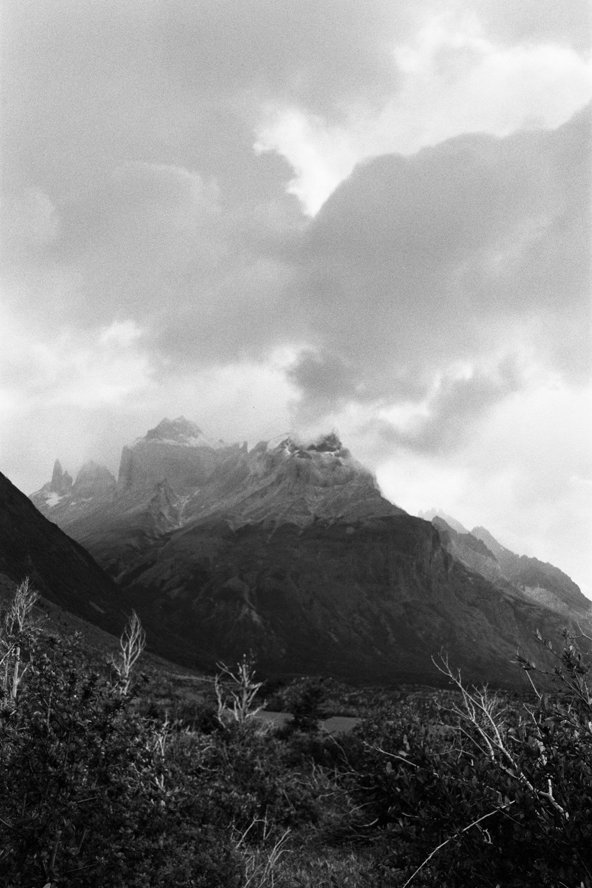 Black-and-white mountain valley with jagged central peak partly shrouded by clouds; foreground shrubs and bare branches.