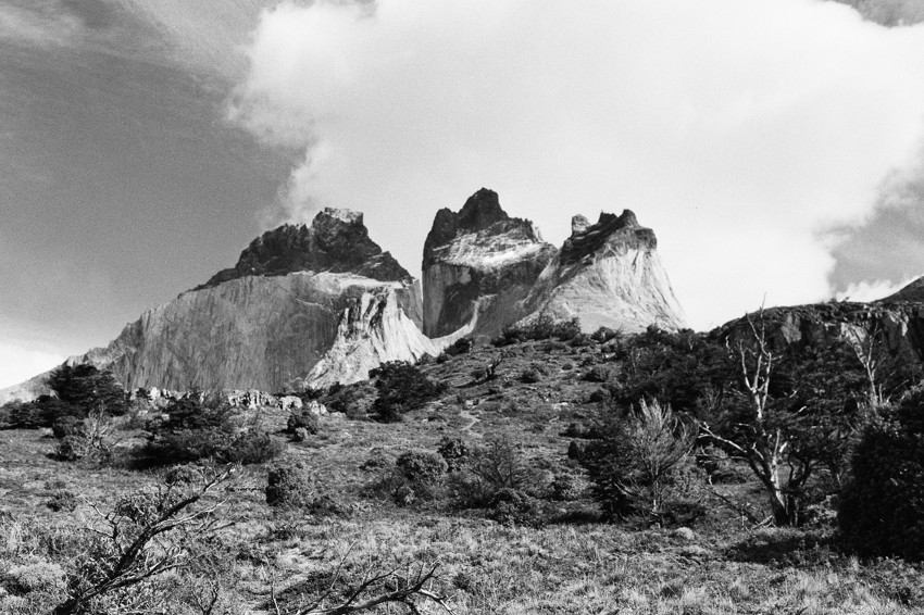 Black-and-white photograph of jagged granite peaks above a shrub-covered hillside with scattered trees and clouds.