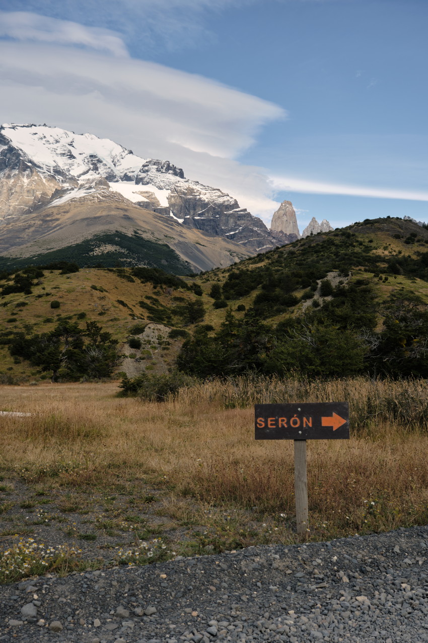 Gravel foreground, wooden sign SERÓN with arrow, grassy hills and snowy mountain peaks under blue sky.