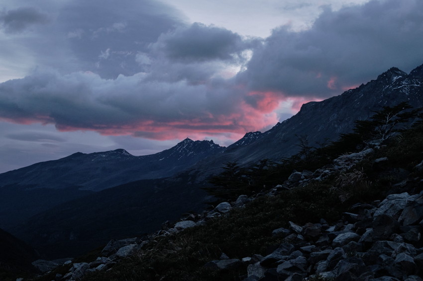 Rocky foreground slope leading to jagged, snow-dusted mountain peaks beneath heavy clouds and pink-tinged sky.
