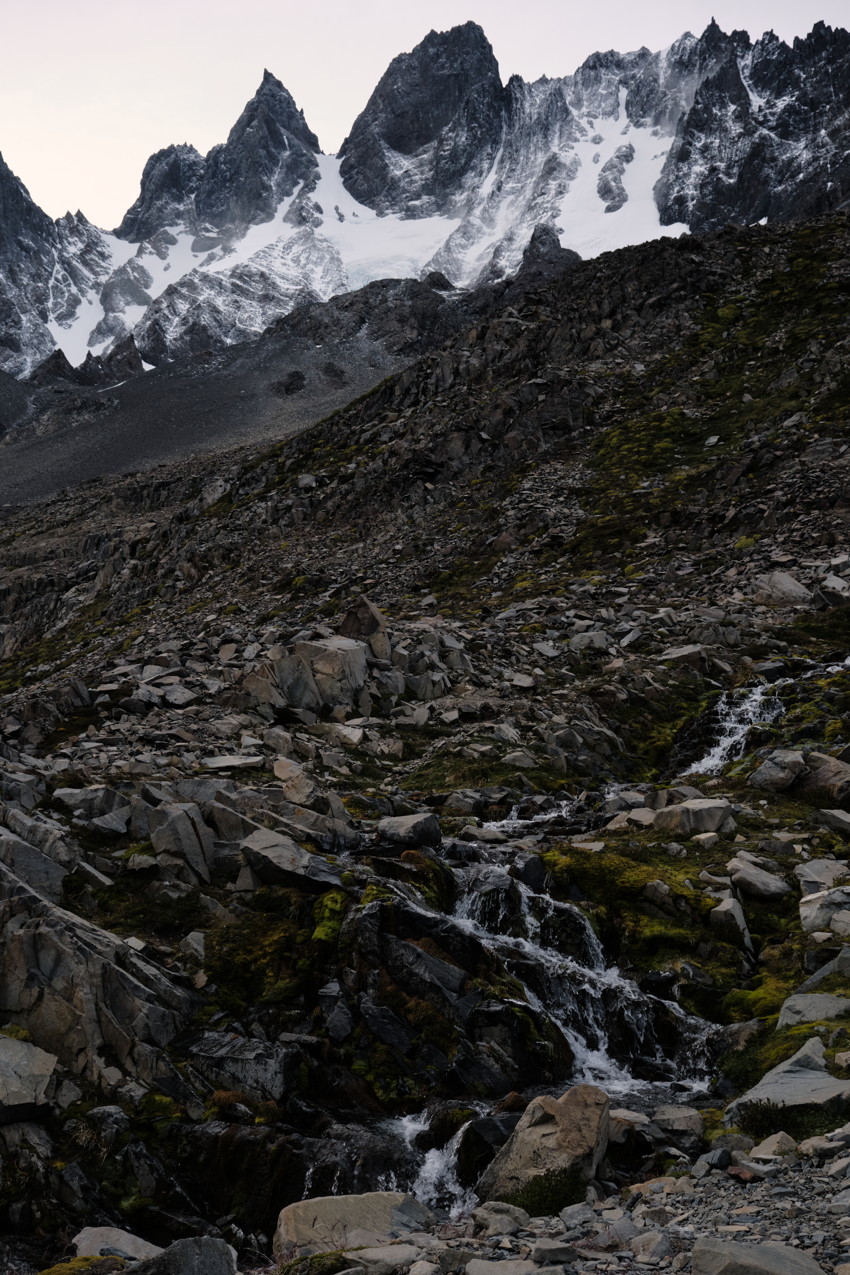 Jagged snow-covered mountain peaks above a rocky scree slope with a small cascading stream over mossy boulders.