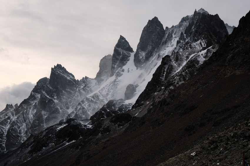 Jagged snow-dusted mountain peaks with steep rocky slopes under overcast sky, mist in the ridges.