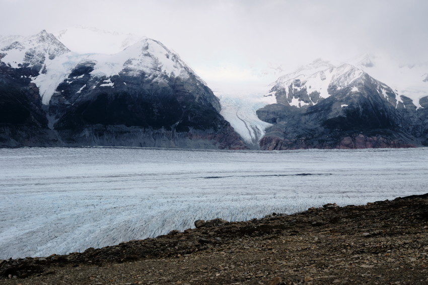 Wide view of glacier filling valley between snow-capped mountains, rocky foreground under overcast sky.