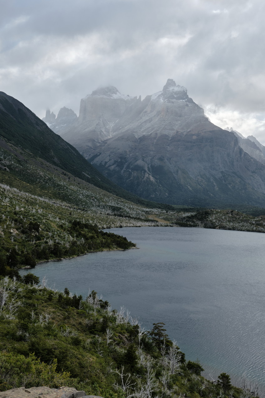 Calm alpine lake in foreground, grassy slope left, rugged snow-dusted mountains under cloudy sky.