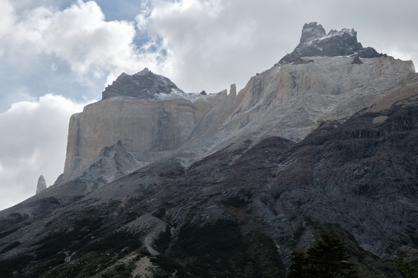 Wide view of steep granite cliffs and jagged peaks above dark, rocky slopes under cloudy sky.