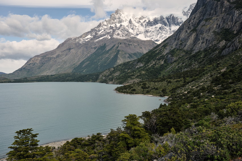 Mountain lake bordered by forest, rocky cliffs on right and snow-capped peaks beneath cloudy sky.