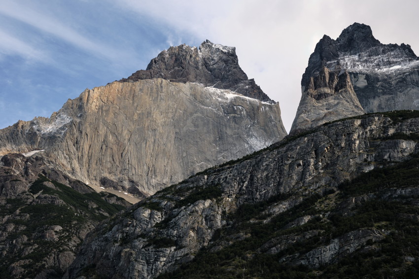 Wide view of steep granite cliff and two jagged mountain peaks above rocky foreground with low vegetation.