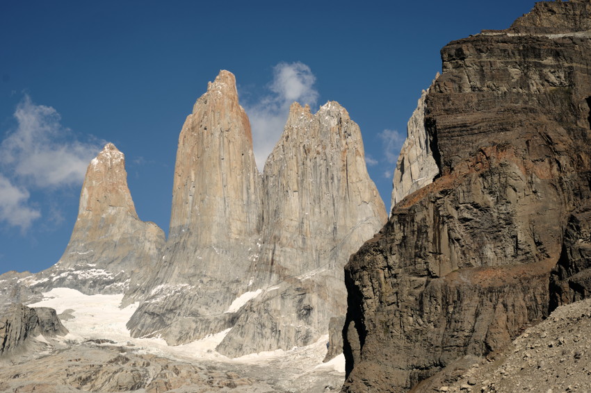 Three steep granite peaks with snow patches under blue sky, dark sheer cliff face at right foreground.