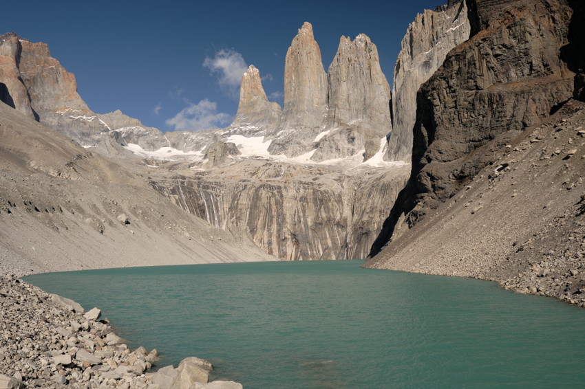 Turquoise glacial lake surrounded by steep rocky moraine with three prominent granite towers and clear blue sky.