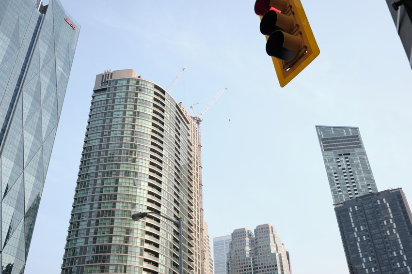 Low-angle view of glass high-rises with construction cranes and a yellow traffic light against clear sky.