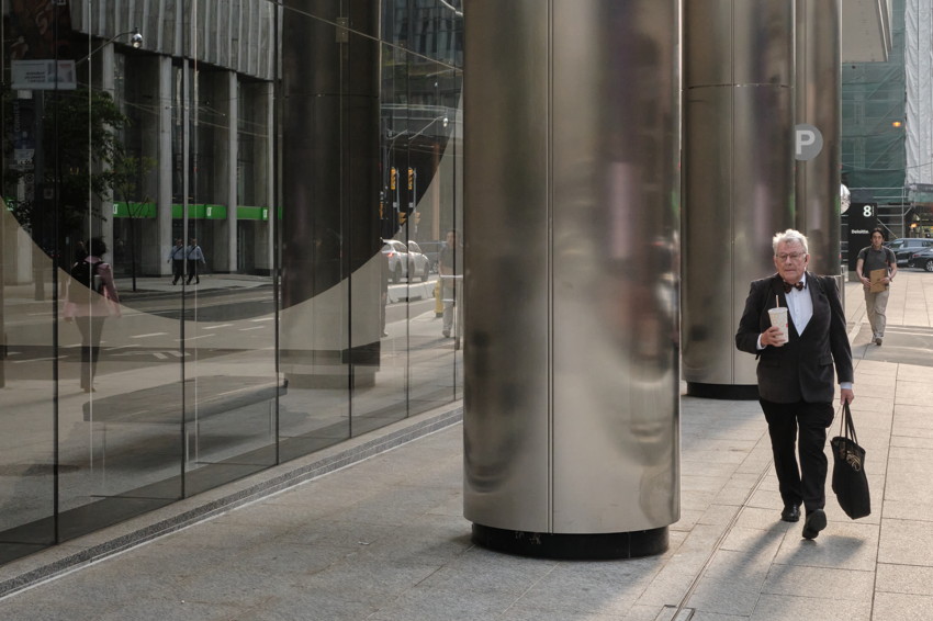 Elderly man in tuxedo holding coffee and bag walking beside large polished metal columns and reflective glass facade.