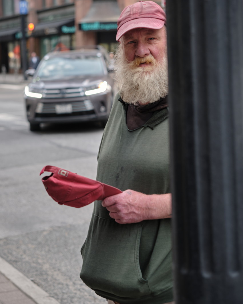 Bearded man in green hoodie and pink cap, holding red hat on urban street, car headlights in background.