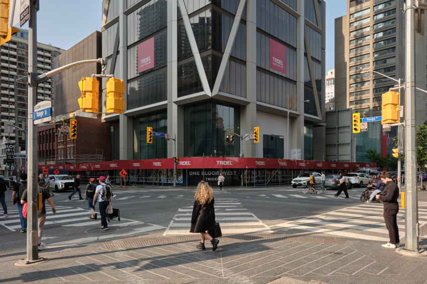 City intersection with glass high-rise, red construction hoarding, traffic lights, and pedestrians crossing toward camera.