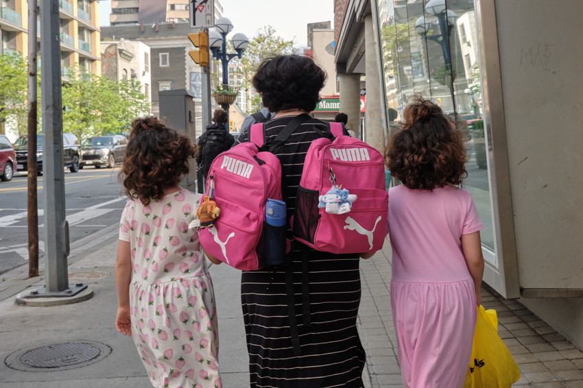 Rear view of three walking on sidewalk: adult with two pink Puma backpacks, two girls in pink dresses.