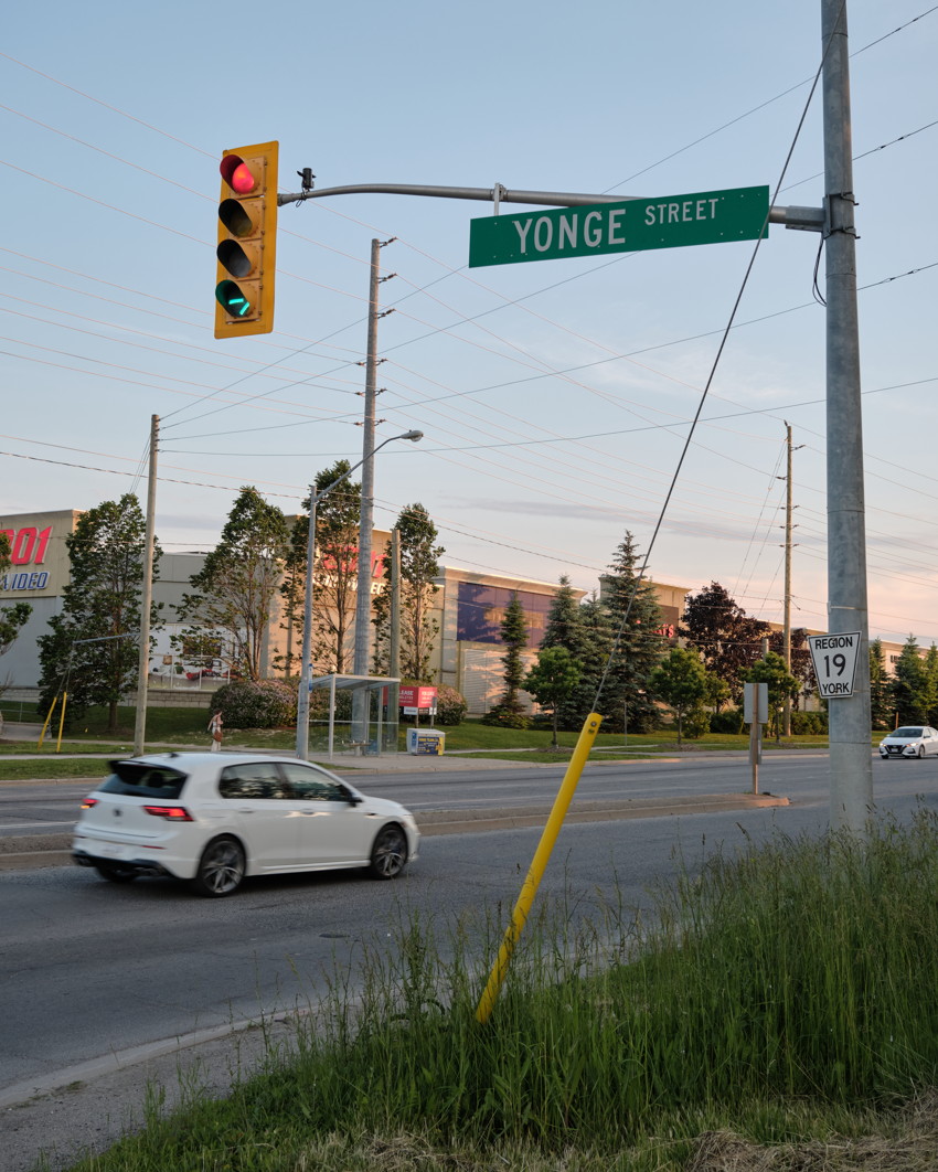 Yonge Street intersection with overhead sign, traffic light, white car on road, utility poles and retail buildings beyond.