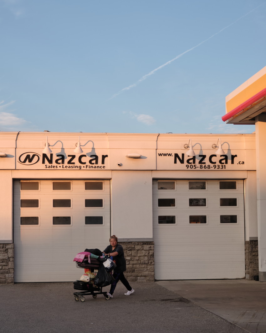 Person pushing a loaded shopping cart past two closed garage doors under 'Nazcar' storefront sign.