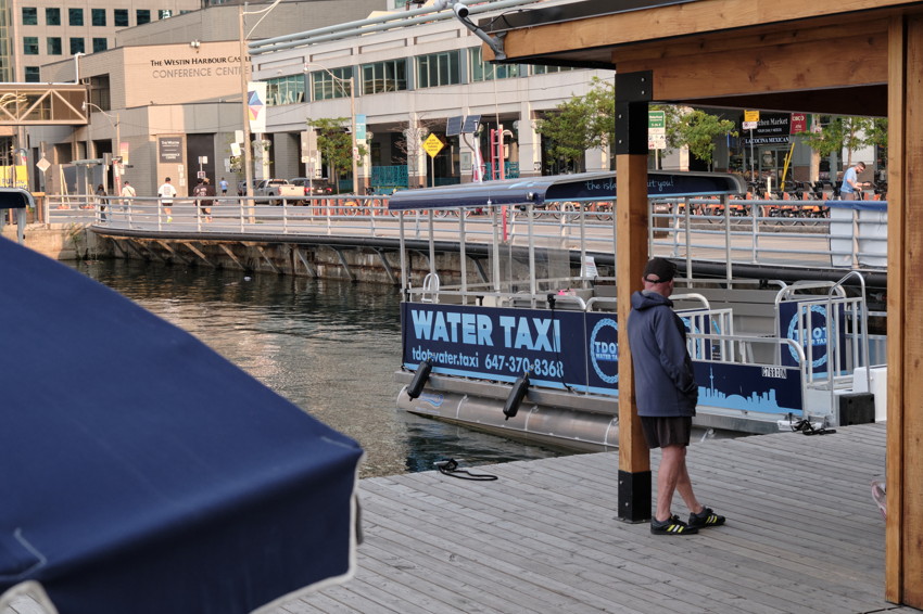 Man stands under wooden shelter on dock beside a docked WATER TAXI boat, water and city buildings behind.