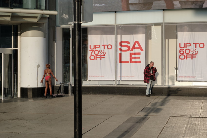 Storefront with large sale posters; person in red swimwear left, person in red jacket right, pole in foreground.