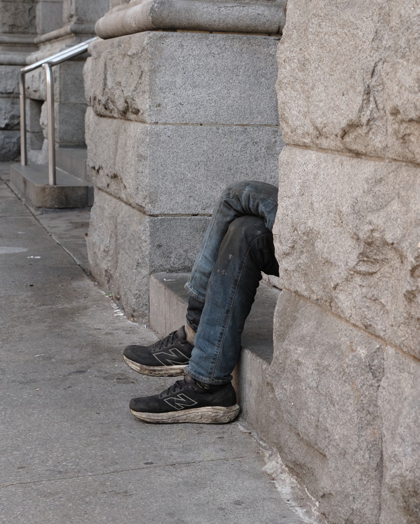 Legs in blue jeans and black New Balance sneakers sitting in a stone building recess beside a concrete sidewalk.