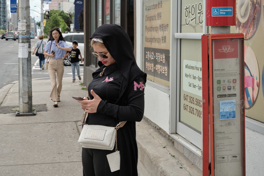 Woman in black hooded jacket checking phone on city sidewalk beside storefront and red transit information kiosk.