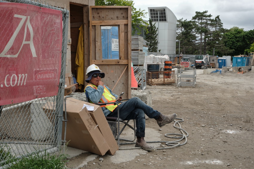 Worker in hard hat and reflective vest sitting on folding chair beside plywood shed at construction site.