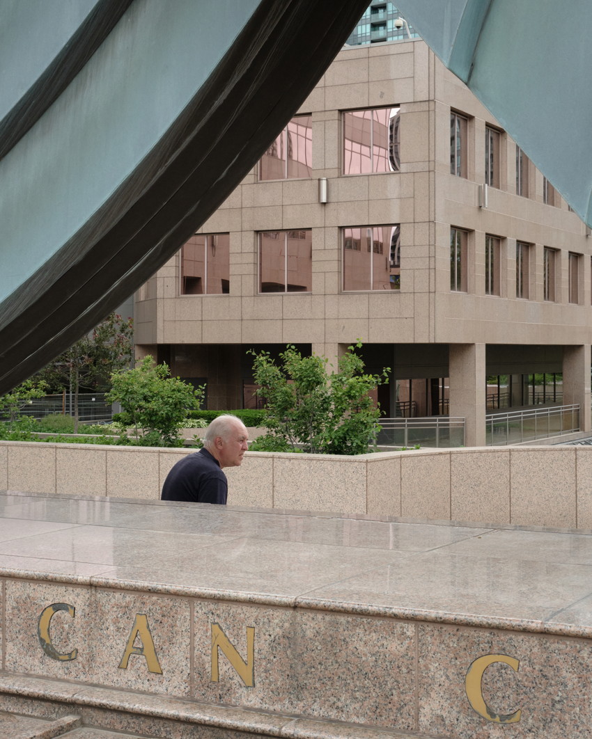 Bald older man walking along marble plaza, midground planters and modern office building framed by draped metal sculpture.