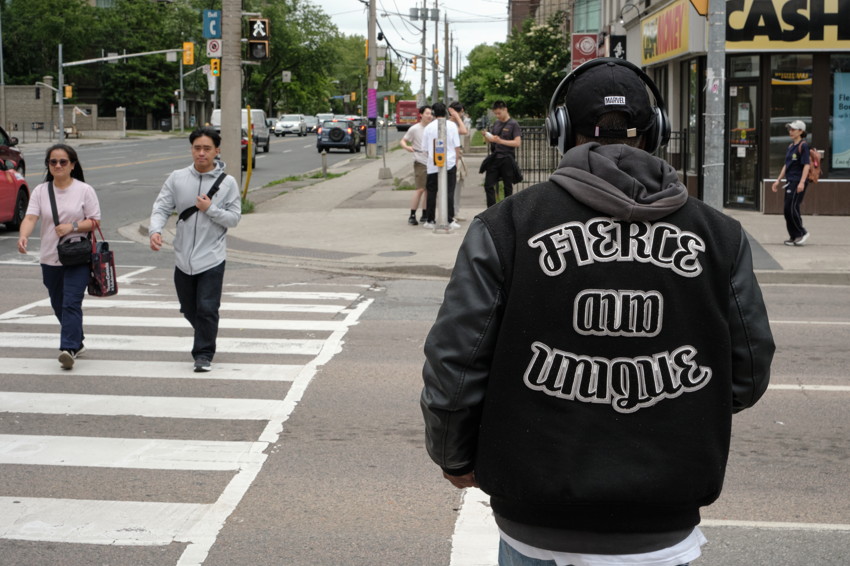 Rear view of person wearing black jacket with 'FIERCE AND UNIQUE' text, standing at city crosswalk.