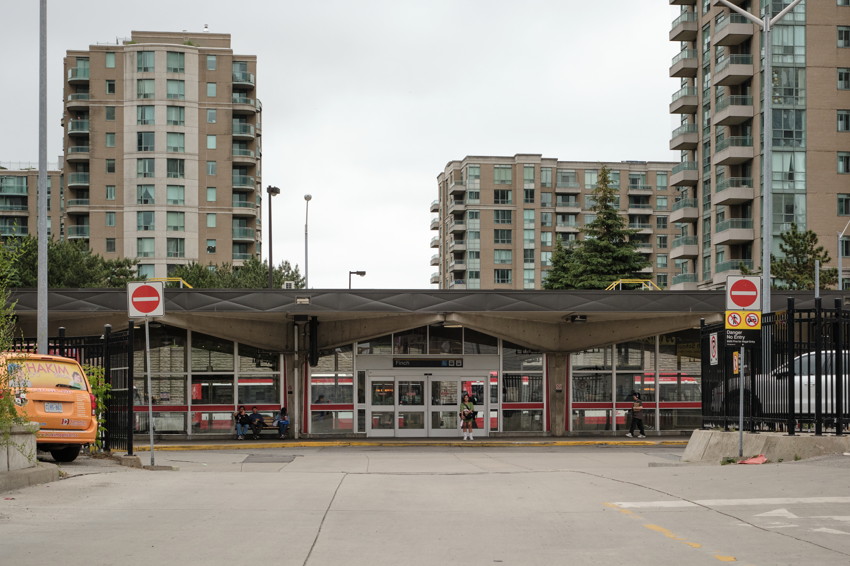 Front view of transit station entrance with angled canopy, apartment towers behind, taxi left, pedestrian near doors.