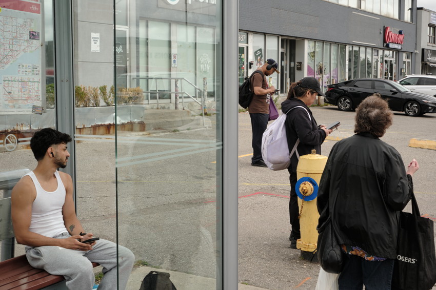 Bus stop scene: man in white tank top seated inside shelter; three people standing by yellow fire hydrant, storefronts.