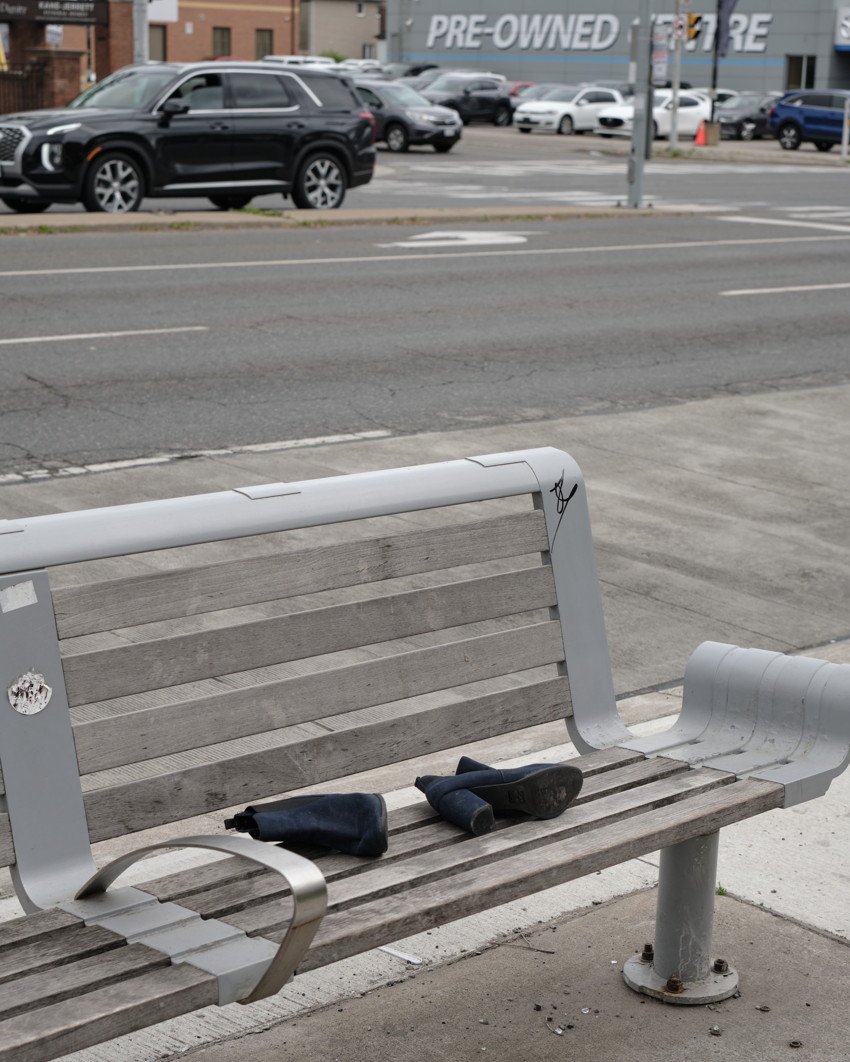 Empty wooden and metal bench with a pair of dark boots placed on the seat; street and parked cars behind.