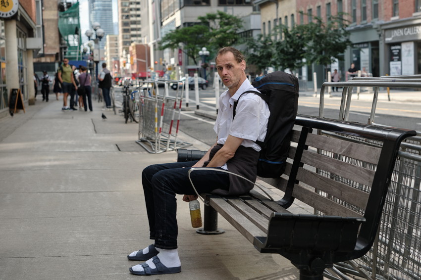 Person sitting on a bench on a city sidewalk with backpack, holding a bottle; socks and sandals visible.