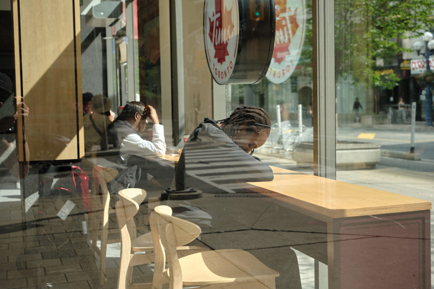 Window view of cafe interior with three people seated at a wooden counter, reflections visible on glass.
