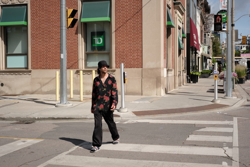 Person wearing black bucket hat and floral shirt walking across crosswalk by brick building with green awnings.