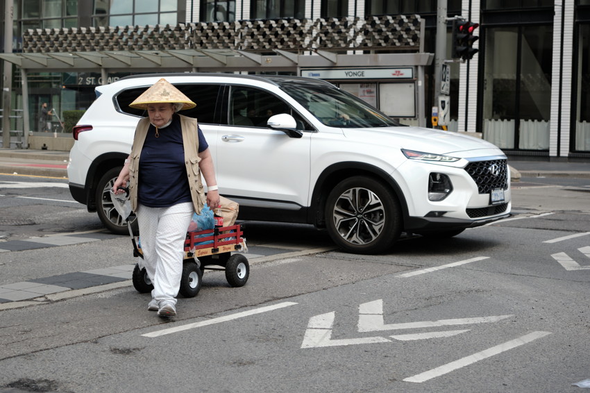 Person wearing conical hat pulls small red wagon across city intersection beside a white SUV.