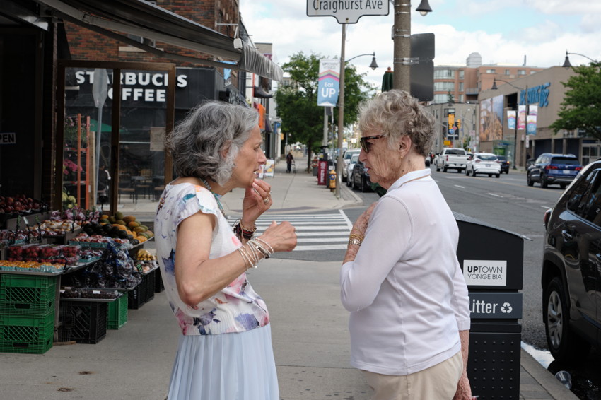 Two older women talking on a city sidewalk beside a produce stand, crosswalk and storefronts visible.