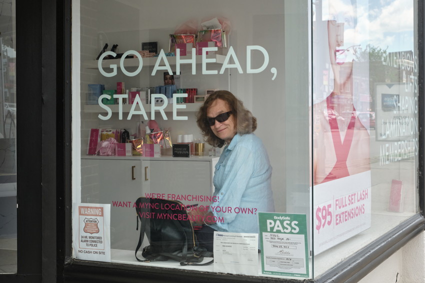 Woman in sunglasses seated behind salon storefront glass with 'GO AHEAD, STARE' lettering and beauty products.