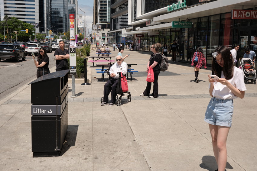 City sidewalk with pedestrians: woman on phone at right, man in wheelchair center, litter bin at left.