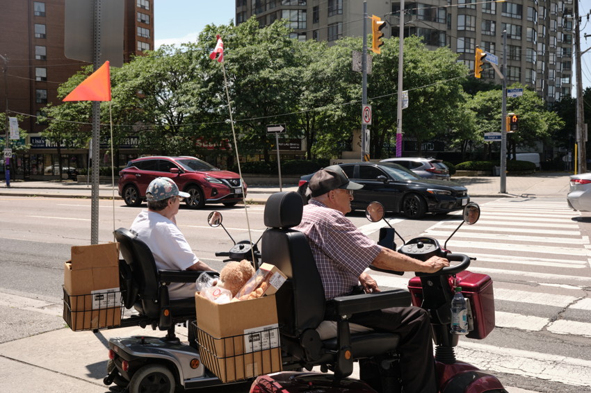 Two people on mobility scooters with grocery baskets and flags, stopped at urban crosswalk.