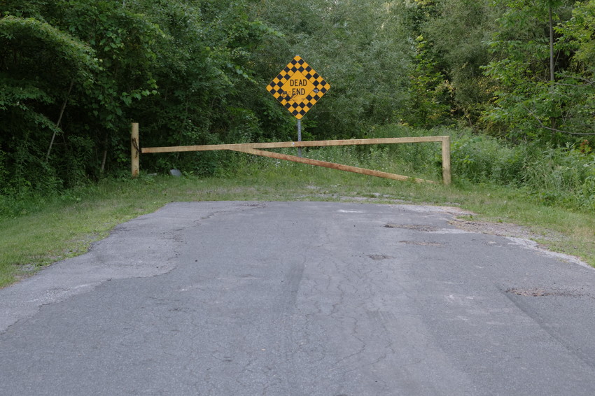 Paved road ending at wooden barrier and yellow checkered 'DEAD END' sign, dense green trees beyond.
