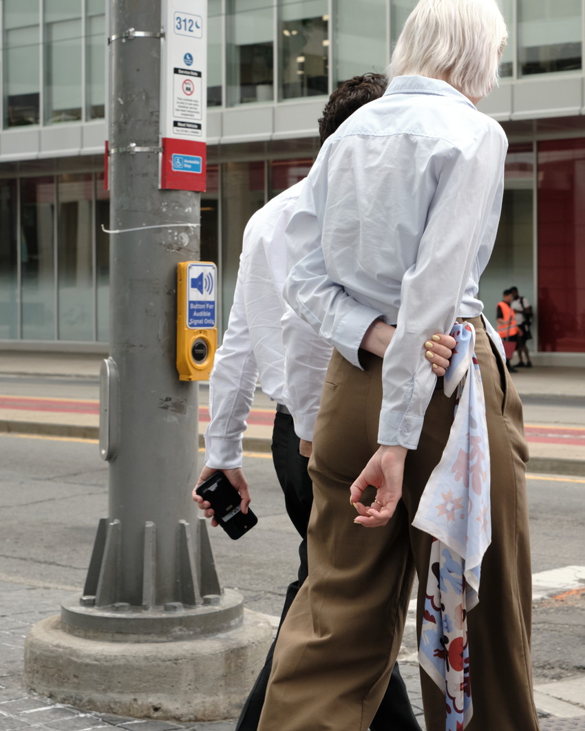 Two people walking at crosswalk, one with white shirt and brown trousers, scarf tucked in pocket, pole and audio button.