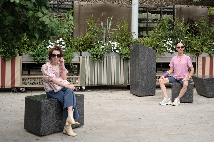 Two people seated on stone blocks in a paved plaza, striped planters with greenery and white flowers behind them.
