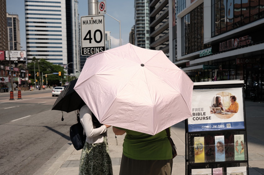 Two people seen from behind under a large pink umbrella on a city sidewalk beside a Bible-course rack.