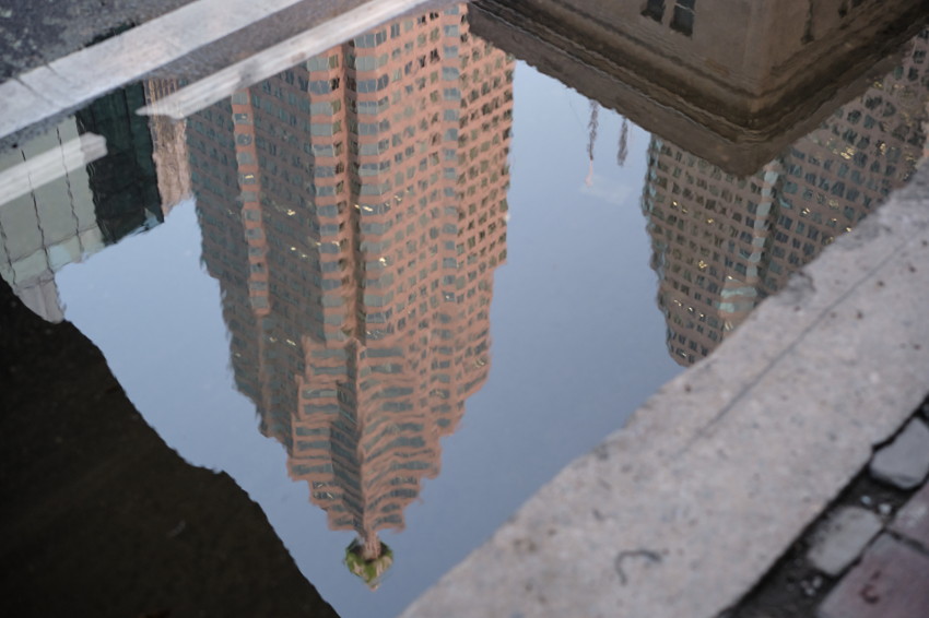 Reflection of tall red-brick skyscraper in a street puddle, bordered by concrete curb and pavement.
