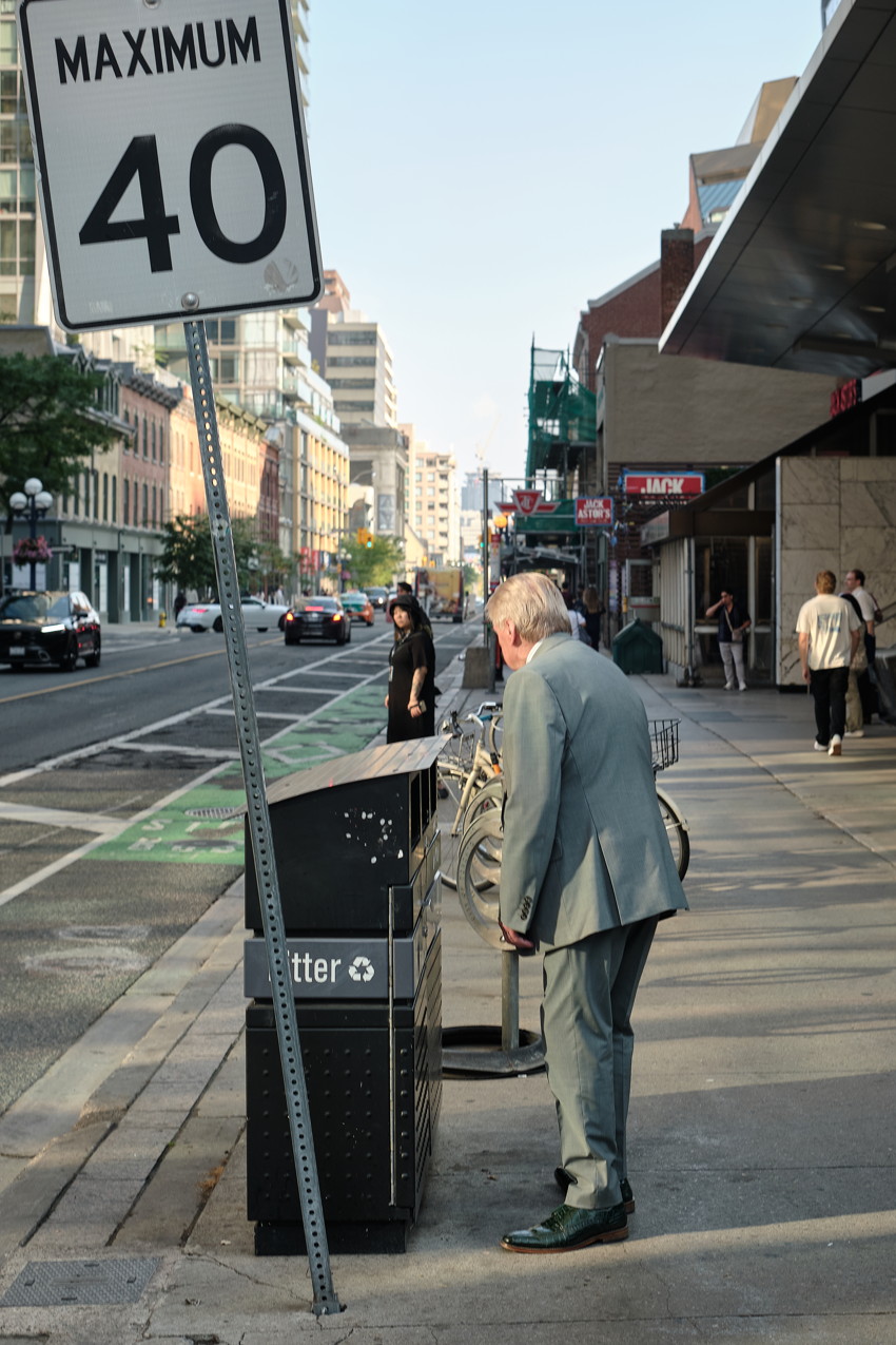 Man in suit leaning into sidewalk recycling bin under 'Maximum 40' sign on urban street.