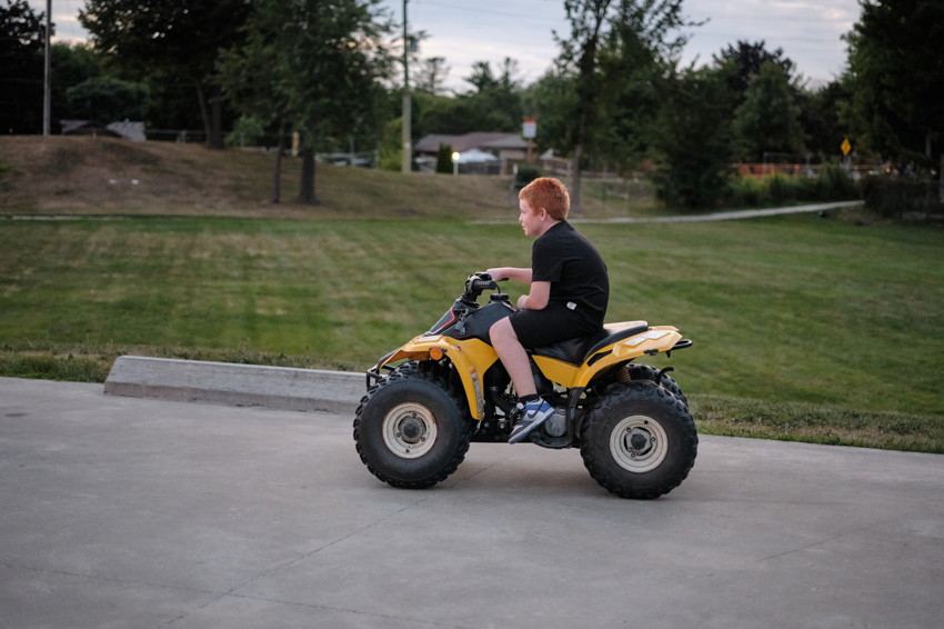 Red-haired boy riding yellow ATV on paved path beside grassy park, profile view facing right.