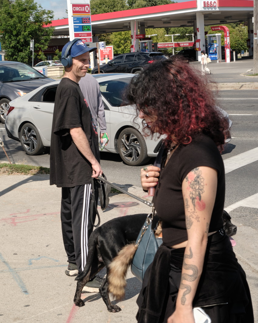 Street scene: woman with tattoos in front, man with headphones holding leash and black dog; gas station background.